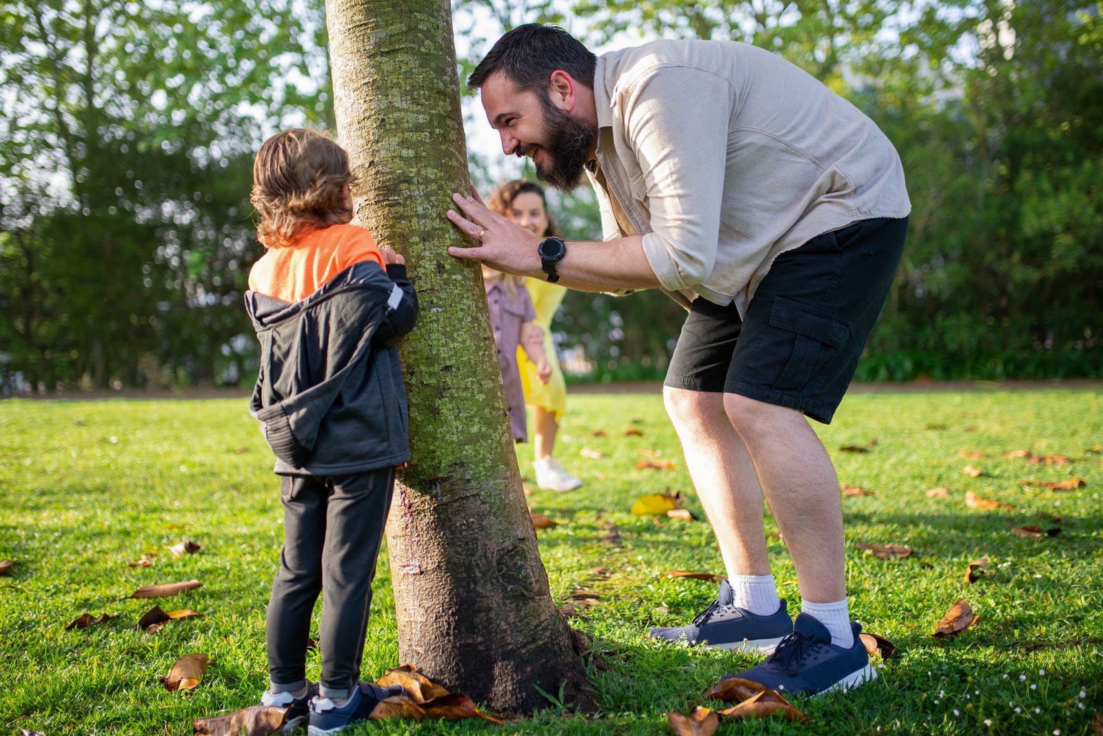 A father and children enjoy playing hide and seek around a tree in a sunny park.
